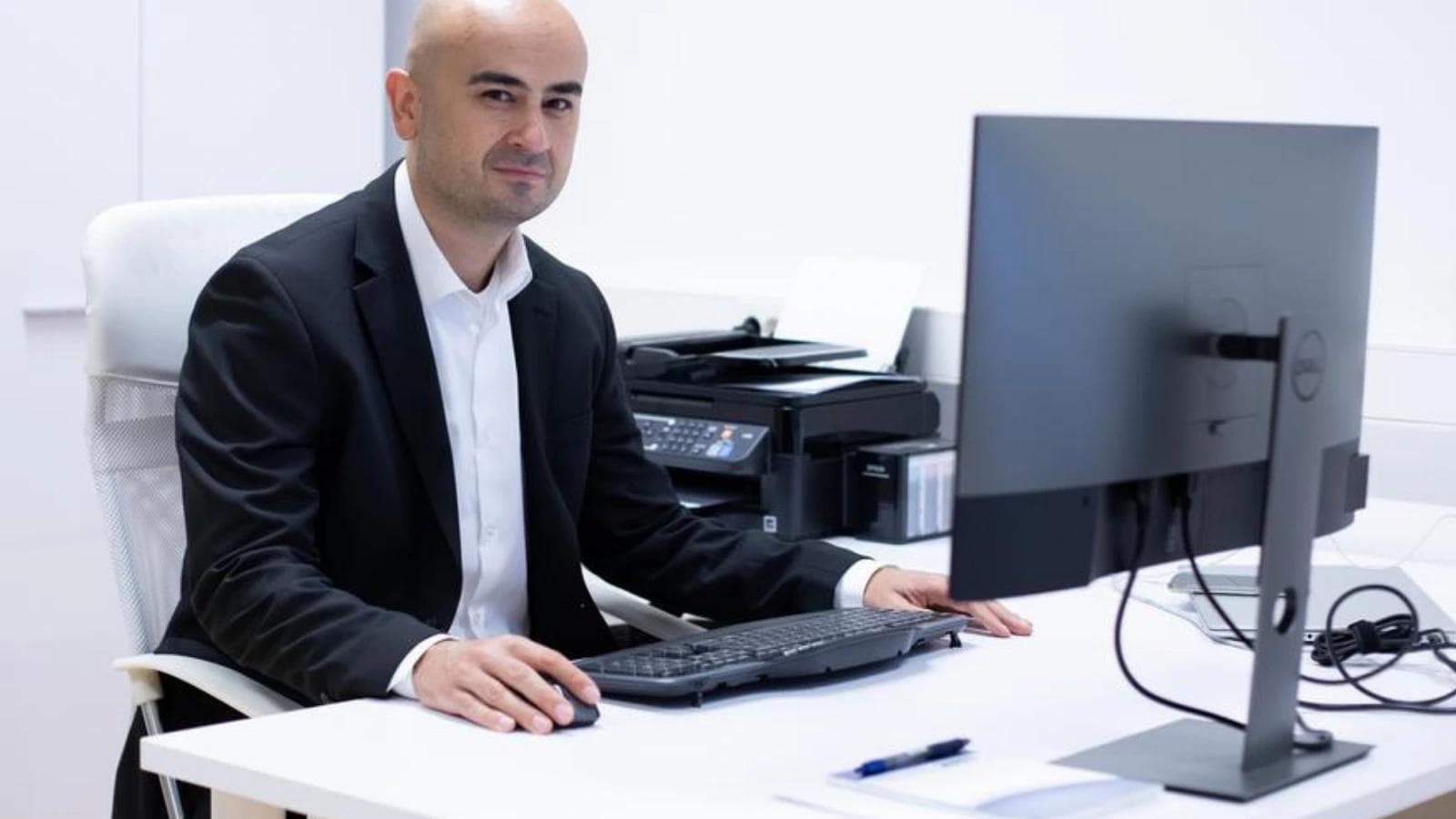 A businessman sitting at his office desk with a computer and printer, smiling at the camera.