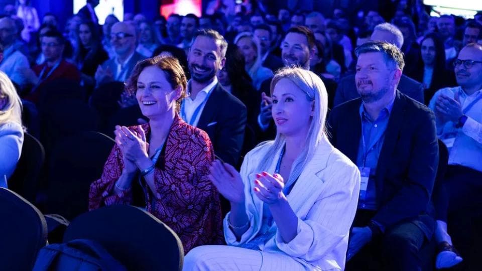 Two woman at an conference clapping surrounded by other people in the audience.